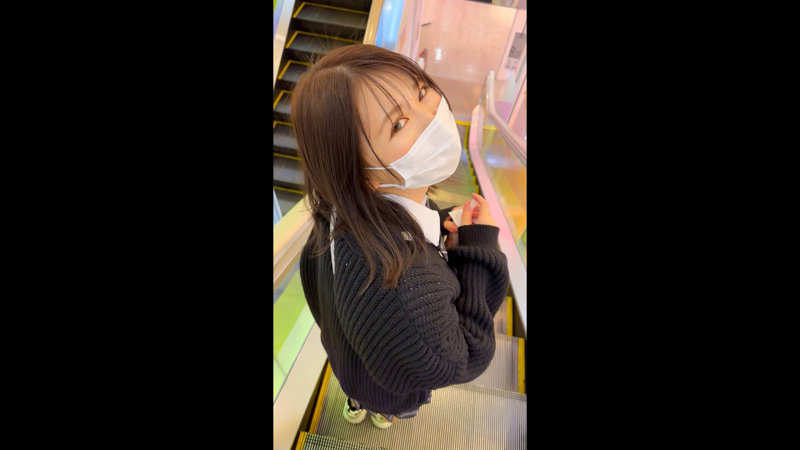 エスカレーターで振り返る、白シャツに黒カーディガンとマスク姿の女性の日常感あるサンプル画像