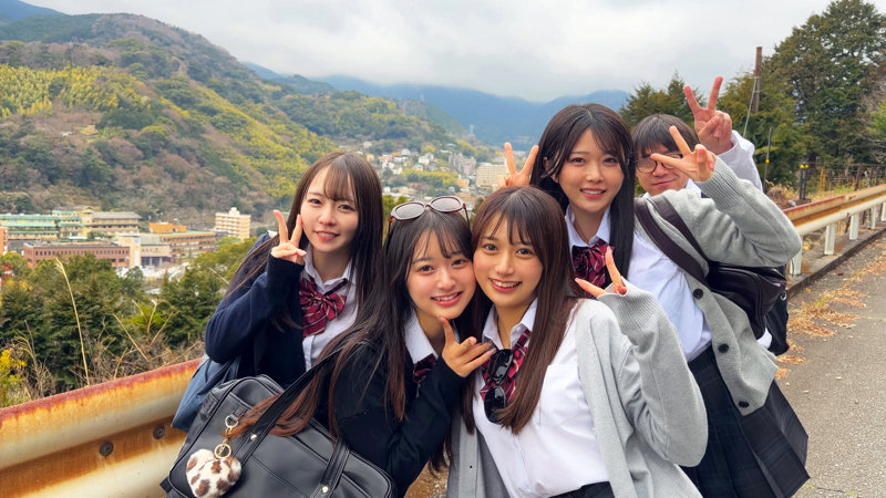 山の景色を背景に制服姿の4人と男性1人が屋外でピースサインを向ける、修学旅行ロケの集合カット