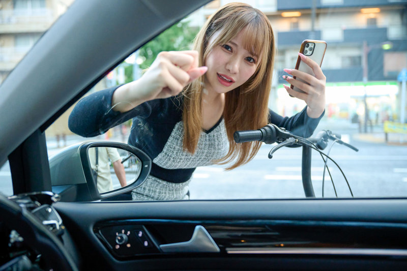 車内から見た窓越しに、自転車のそばに立つ女性がスマートフォンを向ける日常的な場面。